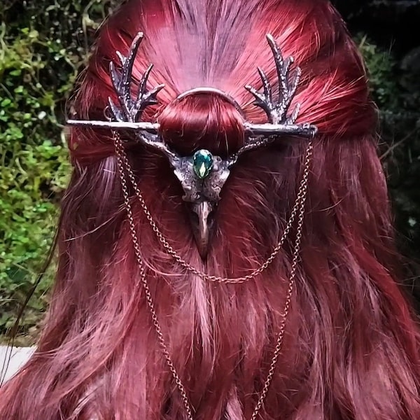 Puede incluir: Un accesorio para el cabello plateado con detalles de cuernos y un diseño central de cráneo de ave con una gema verde. Delicadas cadenas cuelgan del cráneo. El accesorio se coloca sobre cabello rojo.