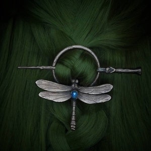 May include: A silver dragonfly hair clip with a blue gemstone in the center. The clip is attached to a silver ring and a silver stick with a decorative end.