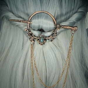 May include: A bronze hair clip with a circular design and a dangling chain. The clip features a decorative floral design with a blue gemstone in the centre.