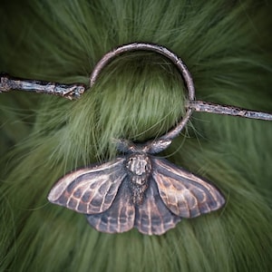 May include: A bronze-colored hair pin with a detailed moth design. The pin has a circular loop and a long, thin, twig-like bar.