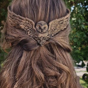 Op de afbeelding: Een bronzen haaraccessoire met een uil met uitgespreide vleugels. Het gedetailleerde ontwerp steekt af tegen een achtergrond van bruin haar, wat een opvallend contrast creëert. Deze haarclip voegt een vleugje door de natuur geïnspireerde elegantie toe.