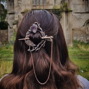 May include: A bronze-coloured hair clip with a bird and branch design, and a purple gemstone. A chain hangs down from the clip. The background shows a blurred view of an old stone building, with a gothic architectural style.