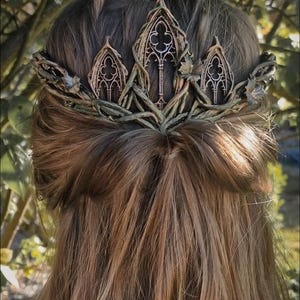 May include: A detailed view of a woman's hair, adorned with an ornate, gold-toned crown. The crown features three arched sections with intricate designs, resembling gothic architecture, and is intertwined with natural-looking vines.