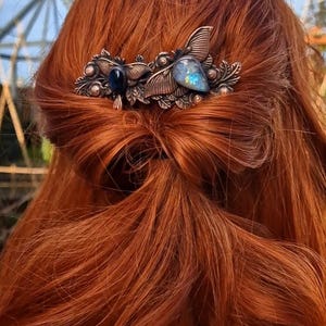 May include: A copper hair clip with a leaf design and two gemstones. The larger gemstone is a blue labradorite and the smaller gemstone is a dark blue stone. The clip is in the hair of a person with long, red hair.