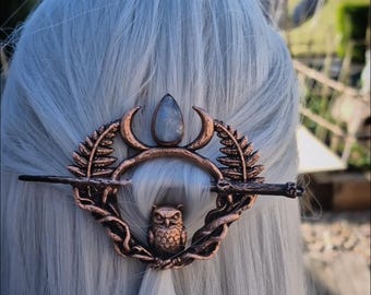 May include: Copper-colored hair barrette featuring an owl, crescent moons, and a central gemstone. The barrette is set against a backdrop of gray hair, with a blurred outdoor scene in the background. A unique hair accessory.
