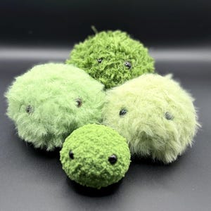 May include: Four round, fuzzy plush creatures in varying shades of green. Each sphere features two small, dark, bead-like eyes embedded within the soft, textured material. The collection includes different textures of green yarn or faux fur.