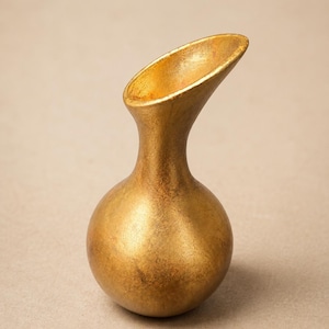 May include: A small, bulbous vase with a metallic gold finish. The vessel features a rounded base that tapers into a slender neck, ending in an elongated, slanted opening. The surface has a textured, slightly weathered appearance with golden tones.