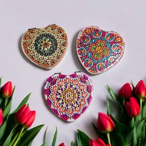 May include: Three heart-shaped compact mirrors with colorful mandala designs made with rhinestones. The mirrors are surrounded by red tulips.