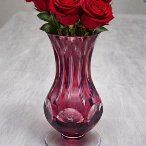 May include: A bouquet of six vibrant red roses in a deep pink glass vase. The vase has a flared top, a bulbous midsection with circular designs, and a circular base. The roses are in full bloom, with green leaves.