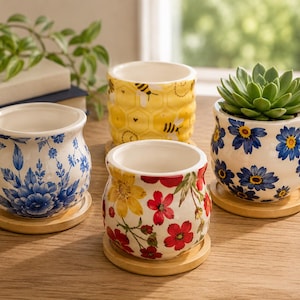 May include: Four small ceramic planters with bamboo saucers. One features a blue floral pattern, one has a yellow honeycomb and bee design, one displays red and yellow flowers, and one contains a green succulent with blue daisy-like flowers.