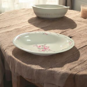 May include: A white ceramic serving bowl with a floral design featuring pink roses and green leaves. The bowl is on a brown table cloth.