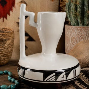 May include: A white ceramic vessel with a wide, rounded base and a narrow neck. The handle features a distinct geometric, stepped design. The lower portion of the vessel is decorated with a black band containing various abstract geometric shapes.