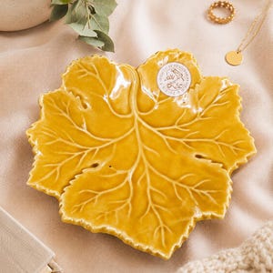 May include: A glossy mustard yellow ceramic dish shaped like a large maple leaf with prominent embossed veins. A small circular white sticker with black text and a floral illustration is adhered to the upper right section of the leaf-shaped dish.