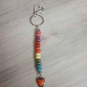 May include: A rainbow beaded keychain with a silver heart charm at the bottom. The keychain has a silver ring and a silver lobster clasp.