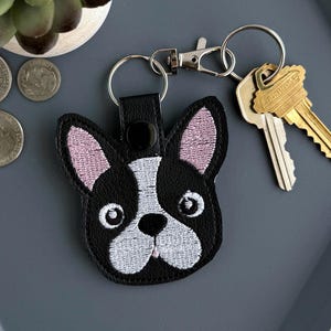 May include: A black and white embroidered French bulldog keychain featuring pink inner ears, attached to a silver metal split ring and lobster clasp. The keychain is accompanied by two brass-colored keys and four assorted silver and copper coins.
