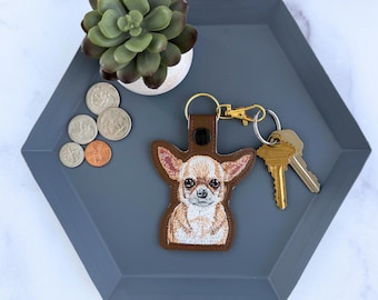 May include: A brown leather-like keychain featuring an embroidered tan and white chihuahua dog portrait. The keychain includes a gold-toned metal lobster clasp and a split ring holding two silver-colored metal keys.