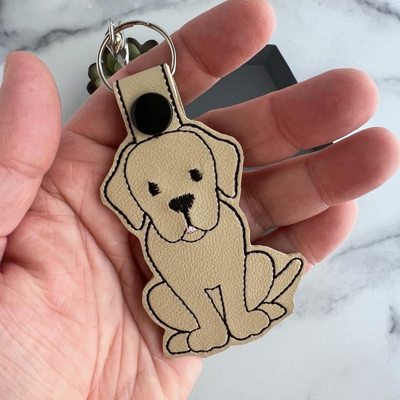 Puede incluir: Un llavero de cuero beige con un contorno negro de un perro Labrador Retriever. El perro est&aacute; sentado con la lengua fuera.