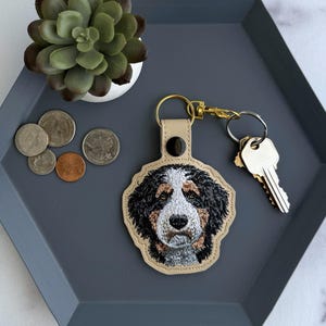 May include: A beige leather keychain featuring an embroidered Bernese Mountain Dog design. The keychain is attached to a gold-coloured clasp and a set of keys. The items are on a grey hexagonal tray with coins and a small succulent.