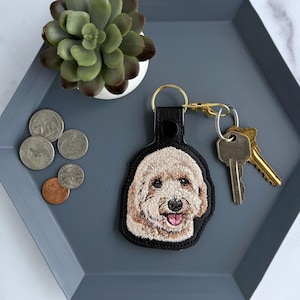 May include: A black leather keychain featuring a detailed embroidered portrait of a light-colored doodle dog with a pink tongue. The keychain includes a gold-toned metal ring and clasp with two silver-toned keys attached to the hardware.