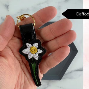 May include: A black leather keychain with a white and yellow embroidered daffodil. The keychain has a gold metal ring.