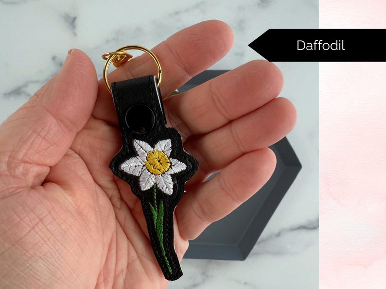 May include: A black leather keychain with a white and yellow embroidered daffodil. The keychain has a gold metal ring.