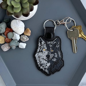 May include: Black and white embroidered wolf head keychain with yellow eyes. The keychain is attached to a silver key ring.