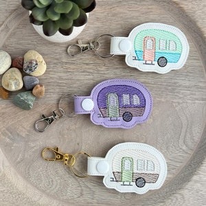 May include: Three keychains featuring a camper design. The keychains are made from white, light purple, and light green fabric. Each keychain has a silver metal keyring and a white button.