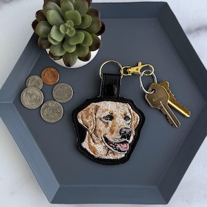 Peut inclure: Un porte-clés en cuir noir avec une image brodée d'un chien golden retriever. Le porte-clés est doté d'un fermoir doré et est attaché à un jeu de clés. Le porte-clés est posé sur un plateau hexagonal gris avec des pièces de monnaie et une plante succulente.