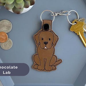 Puede incluir: Llavero de perro labrador chocolate con un dise&ntilde;o de cuero marr&oacute;n. El llavero tiene un anillo de metal plateado y un cierre de metal plateado.