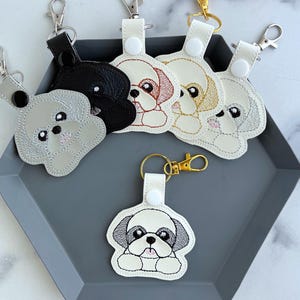 May include: A collection of six dog-shaped keychains featuring embroidered faces in various colors including black, white, gray, and tan. Each keychain has a metal lobster clasp, a snap button closure, and a stitched outline of a puppy head.