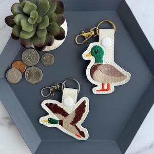 May include: Two duck-shaped keyrings with embroidered details. One shows a duck in flight, the other a standing duck. The ducks have green heads, brown bodies, and orange feet. They are on white leather with gold-coloured hardware.