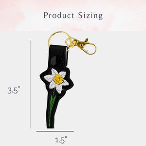 May include: Black leather keychain with a white embroidered flower and a gold key ring. The keychain measures 3.5 inches in length and 1.5 inches in width.