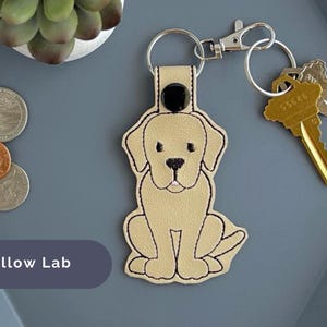Puede incluir: Un llavero de cuero beige con un labrador retriever amarillo bordado. El llavero tiene un bot&oacute;n negro y un anillo de llave plateado.