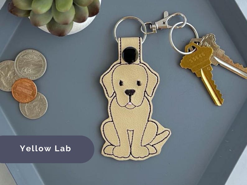 Puede incluir: Un llavero de cuero beige con un labrador retriever amarillo bordado. El llavero tiene un bot&oacute;n negro y un anillo de llave plateado.