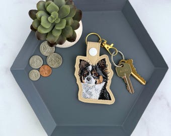 May include: A beige faux leather keychain featuring an embroidered portrait of a tri-color long-haired dog with black, white, and tan fur. The keychain has a gold-tone metal lobster clasp and a split ring holding two silver-tone metal keys.
