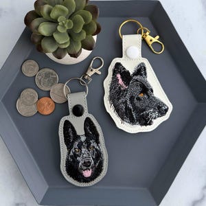 May include: Two keychains with embroidered black German Shepherd dog portraits. One keychain is gray, the other is white. Both have metal clasps. The keychains are on a gray tray with coins and a small succulent plant.