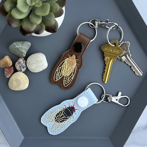 May include: Two cicada-themed keychains, one brown and one light blue, with metal key rings and clasps. The brown keychain has a black button and the light blue keychain has a white button. Keys are also present.