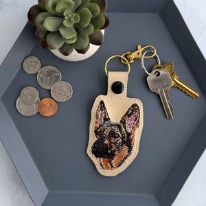 May include: A tan leather keychain featuring an embroidered German Shepherd dog portrait. The keychain has a gold-tone clasp and a black snap closure. Keys are attached to the clasp. The keychain rests on a gray tray with coins and a small succulent.