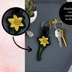 May include: A black leather keychain with a yellow daffodil embroidered on it. The keychain has a gold metal ring and a black metal clasp. The text "Handmade with attention to detail" is visible in the image.