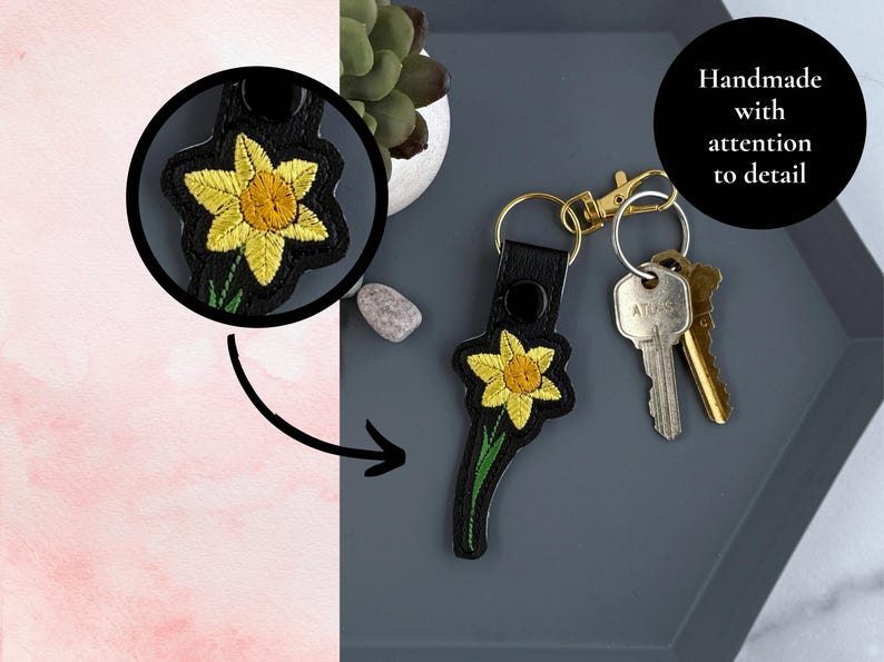 May include: A black leather keychain with a yellow daffodil embroidered on it. The keychain has a gold metal ring and a black metal clasp. The text "Handmade with attention to detail" is visible in the image.