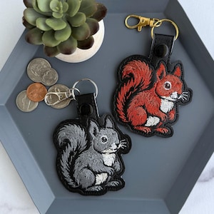 May include: Two embroidered squirrel keyrings, one red and one grey, with black leather straps and gold-tone clasps. The keyrings are on a grey hexagonal tray with a small succulent and coins.