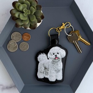 May include: A black leather keychain with an embroidered Bichon Frise dog, tongue showing. The keychain is attached to a gold-coloured clasp and ring, with keys. The keychain rests on a grey tray with coins and a small succulent plant.
