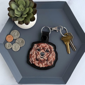 May include: A black leather keychain with a detailed embroidered Cavalier King Charles Spaniel dog face. The keychain is on a gray hexagonal tray with keys, coins, and a small succulent plant in a white pot.
