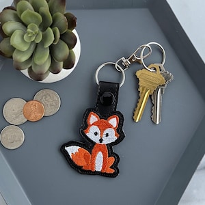 May include: A black leather keychain with a cute orange and white fox design. The keychain has a silver split ring and a silver key ring.