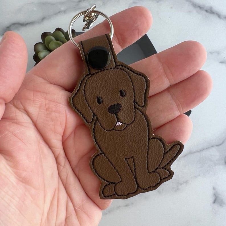 Puede incluir: Llavero de cuero marr&oacute;n con un dise&ntilde;o de labrador retriever marr&oacute;n cosido. El llavero tiene un anillo de llave de metal plateado.