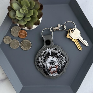 May include: A gray leather keychain featuring an embroidered portrait of a Bernedoodle dog. The keychain is attached to a silver clasp and a set of keys. The keychain rests on a gray hexagonal tray with coins and a small succulent.