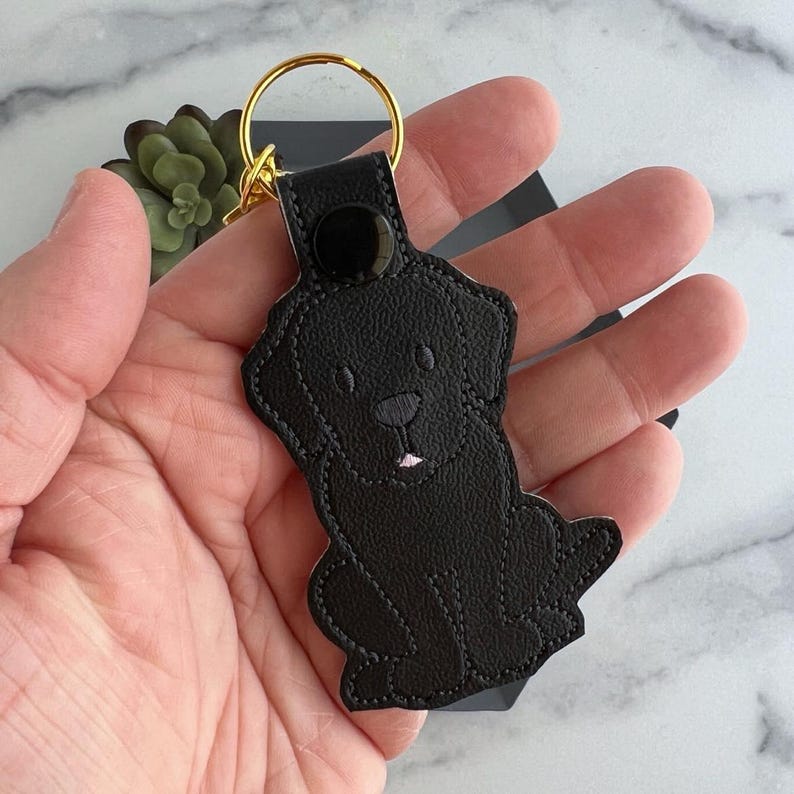 Puede incluir: Llavero de cuero negro con un dise&ntilde;o de labrador retriever negro. El llavero tiene un anillo de metal dorado.