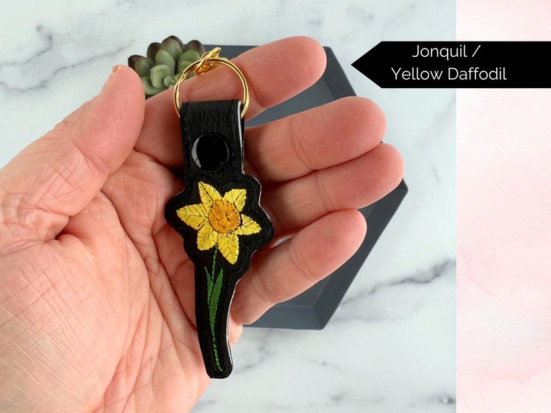 May include: A black leather keychain with a gold key ring and an embroidered yellow daffodil. Jonquil / Yellow Daffodil.