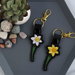 May include: Two black leather keychains with embroidered daffodils. One keychain has a white daffodil and the other has a yellow daffodil.