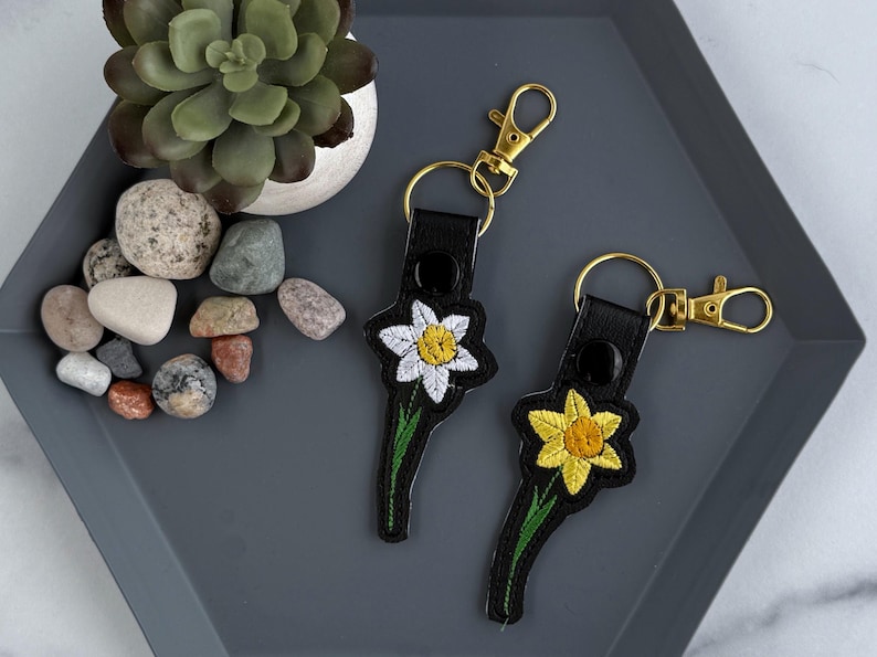 May include: Two black leather keychains with embroidered daffodils. One keychain has a white daffodil and the other has a yellow daffodil.